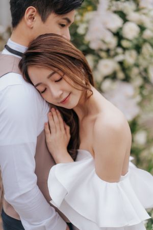 A romantic wedding couple embraces tenderly against a backdrop of white flowers in a dreamy outdoor setting一對浪漫的新婚夫婦在夢幻般的戶外環境中，以白色花朵為背景，深情擁抱