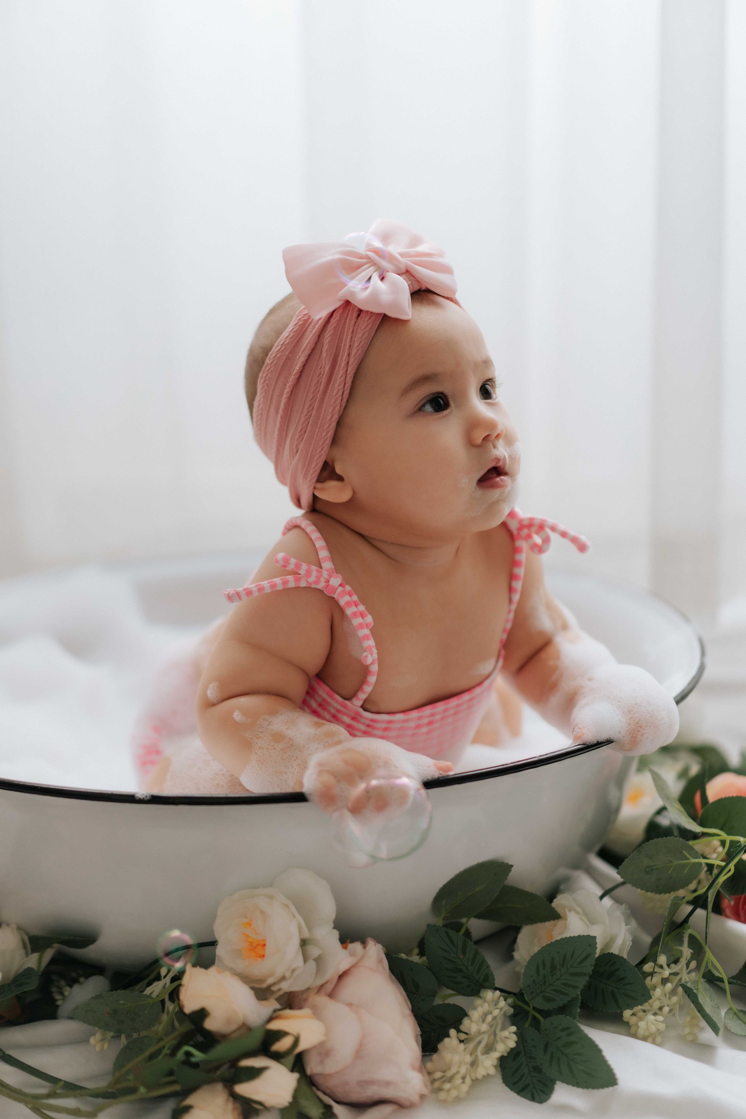 Baby wearing pink headband rests in white ceramic bowl surrounded by delicate flowers and greenery.戴著粉紅色髮帶的嬰兒躺在白色陶瓷碗裡，周圍環繞著嬌嫩的花朵和綠葉。