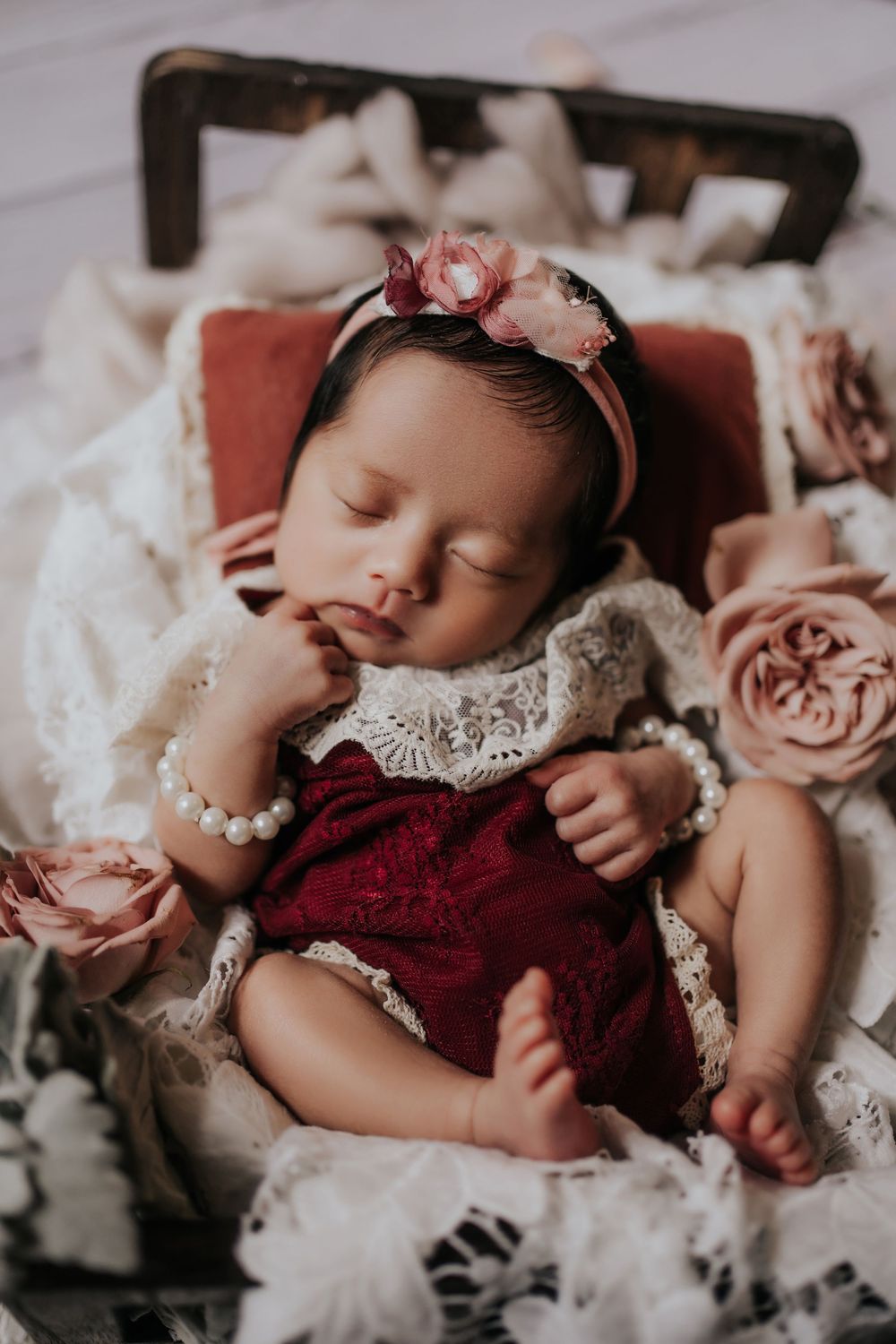 A sleeping baby wearing a burgundy and lace dress lies among soft pink roses and white tulle fabric.一個穿著酒紅色蕾絲連身裙的熟睡嬰兒躺在柔軟的粉紅色玫瑰和白色薄紗中。