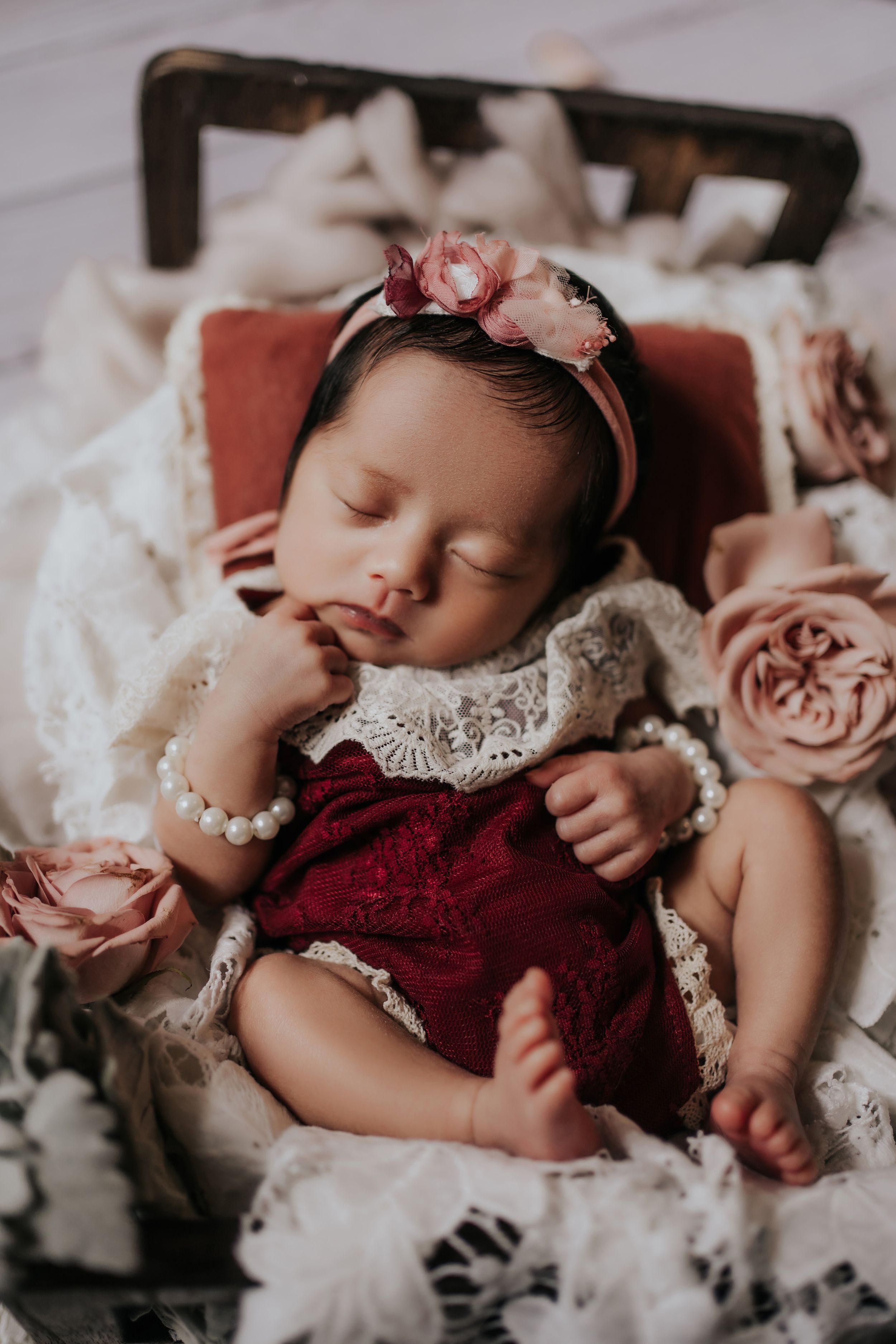 A sleeping baby wearing a burgundy and lace dress lies among soft pink roses and white tulle fabric.一個穿著酒紅色蕾絲連身裙的熟睡嬰兒躺在柔軟的粉紅色玫瑰和白色薄紗中。