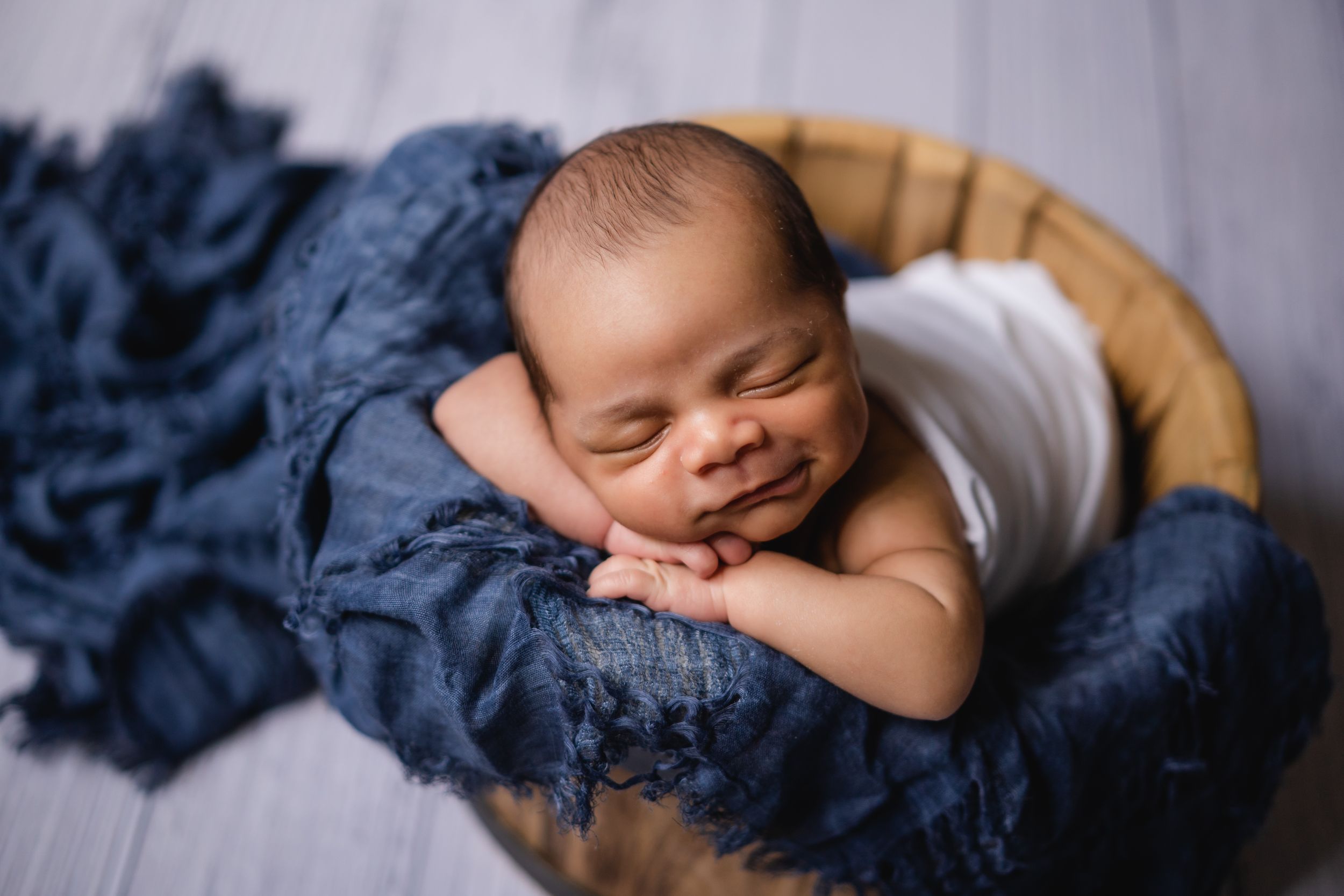 Peaceful sleeping newborn wrapped in white cloth rests on navy blue fabric in wooden basket裹在白布里的新生兒安然入睡，躺在木籃裡的深藍色布料上。