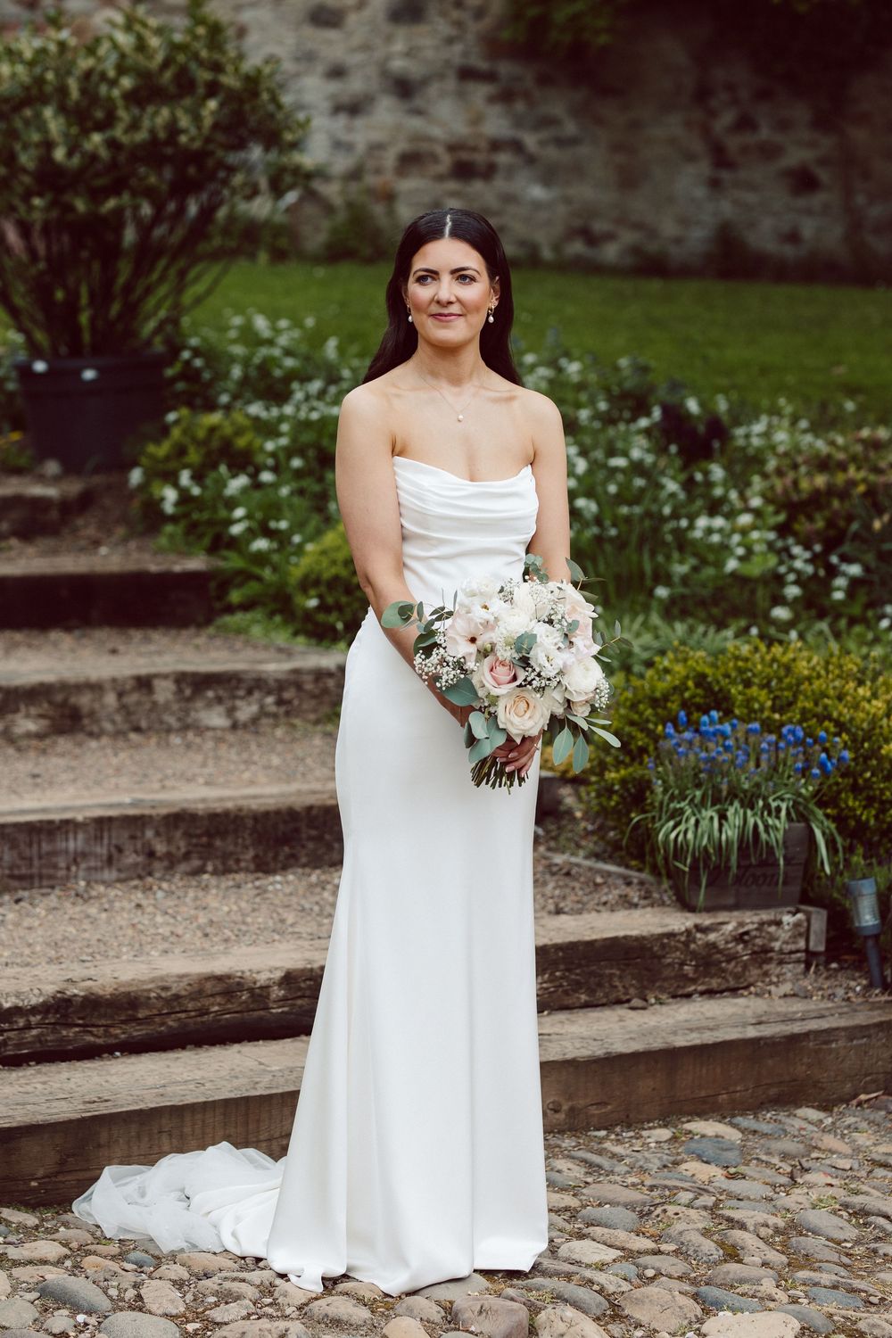 A bride in a white strapless wedding dress holds a pink and white bouquet in a garden setting.