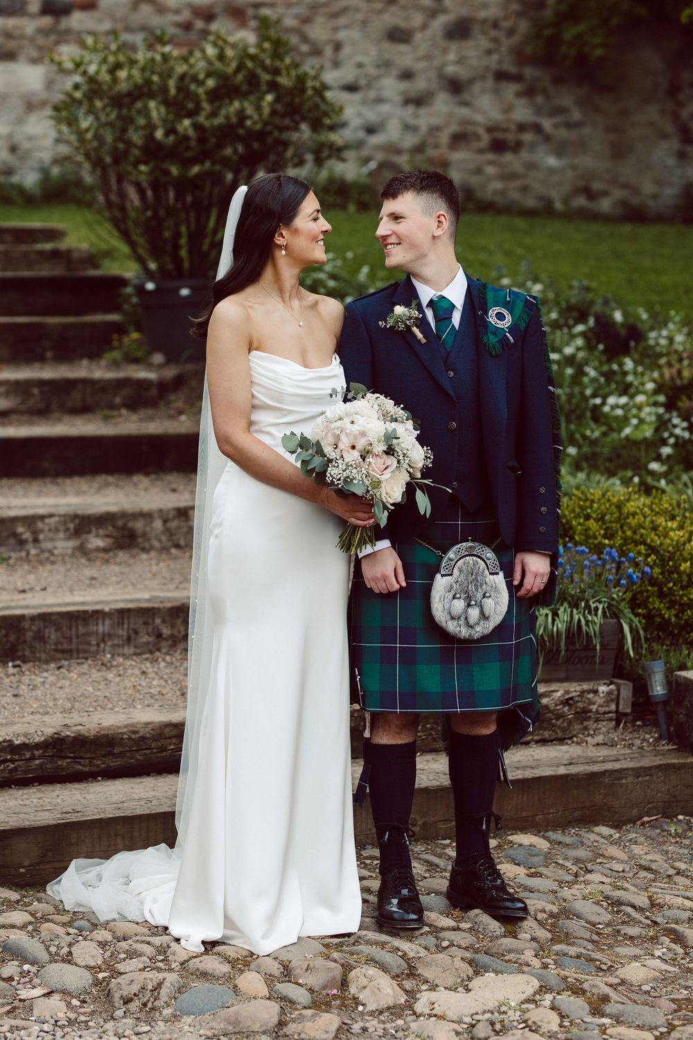 A couple in traditional Scottish wedding attire pose together on a cobblestone path.