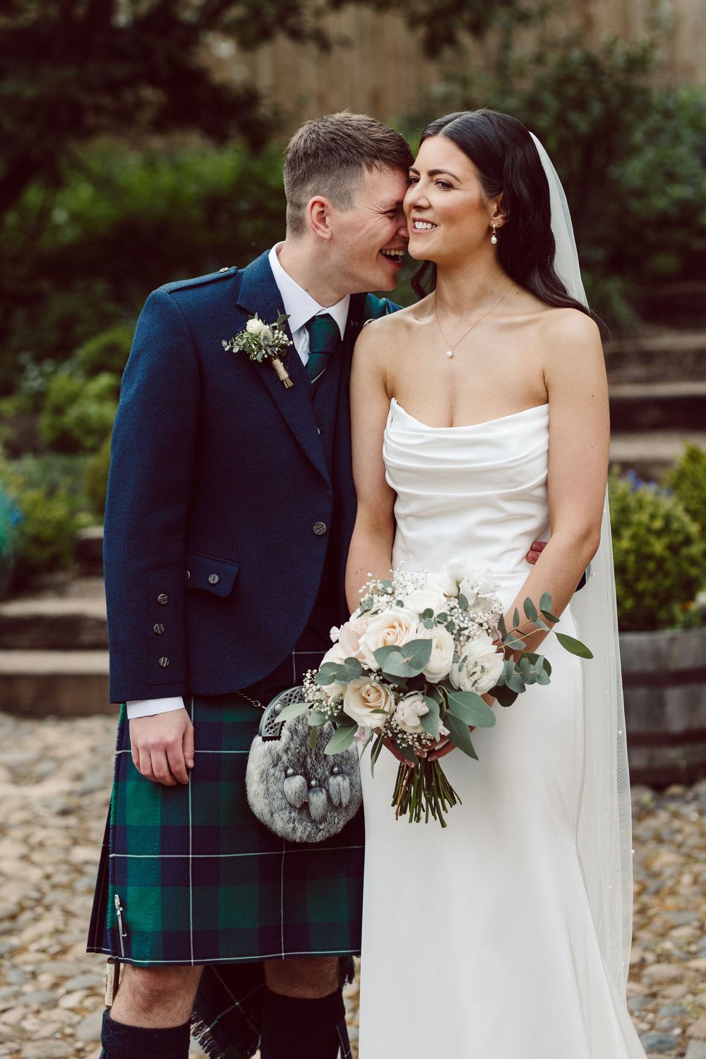 Romantic wedding scene with a couple in traditional Scottish attire embracing outdoors among greenery.