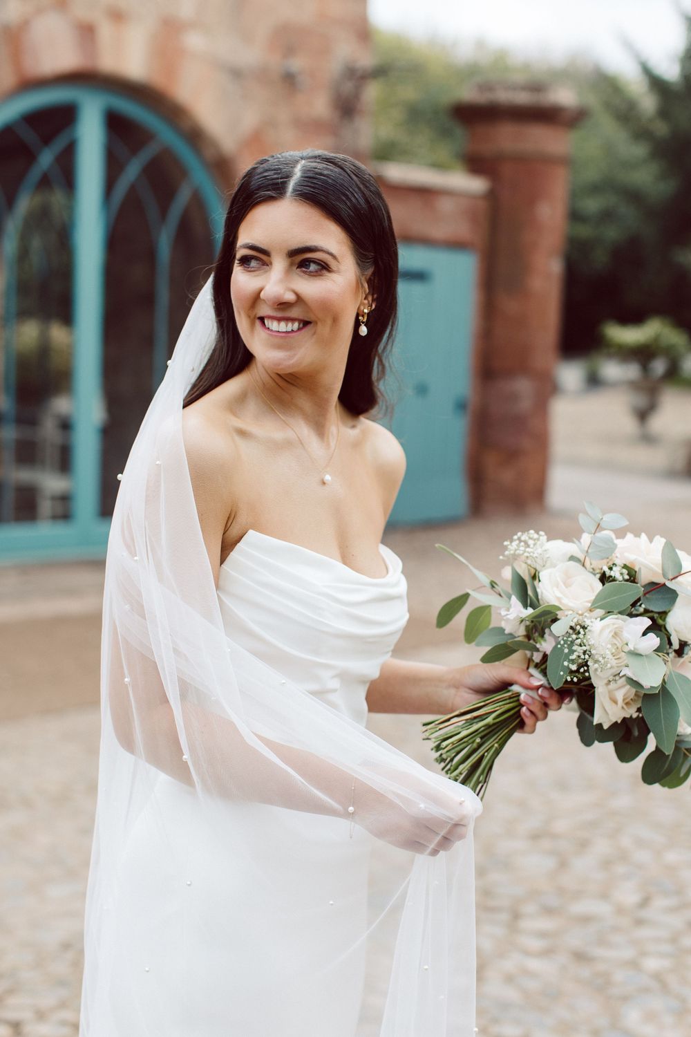 A bride in a white wedding dress and veil holds a romantic bouquet of white flowers and greenery against a rustic backdrop.