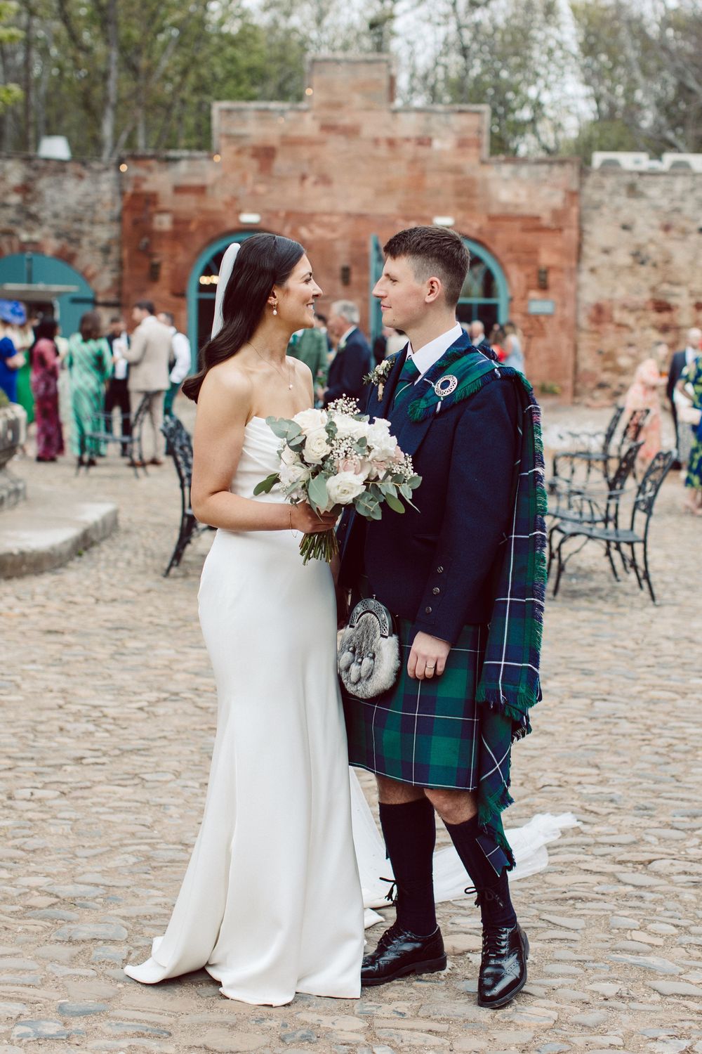 A couple in traditional Scottish wedding attire shares intimate moments on cobblestone grounds.
