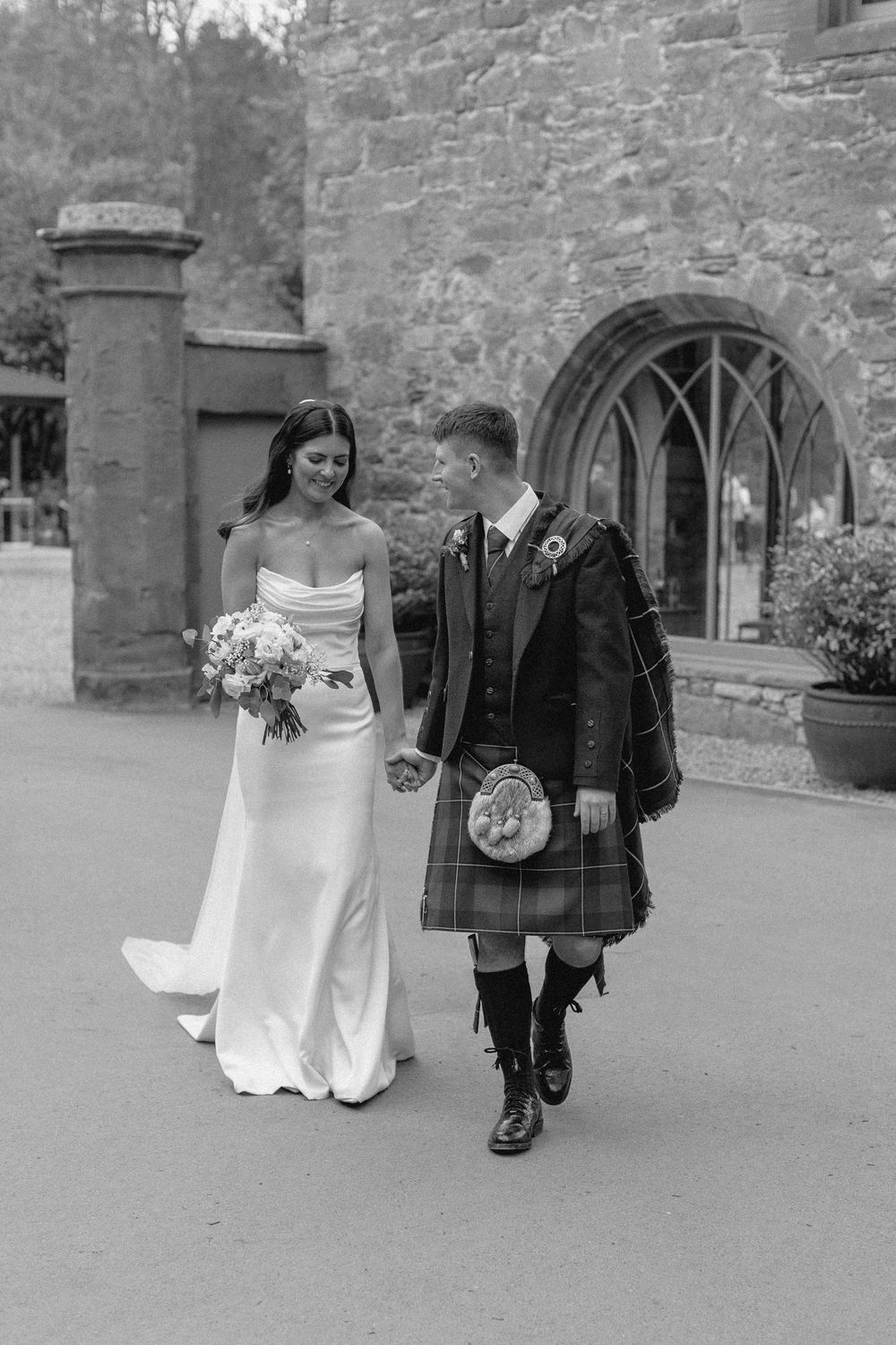 A bride and groom walk together on a stone path outside a historic building in black and white photography.