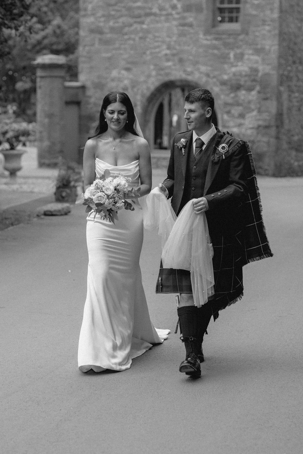 A bride in white dress walks with a young ring bearer in a suit at a stone church wedding ceremony.