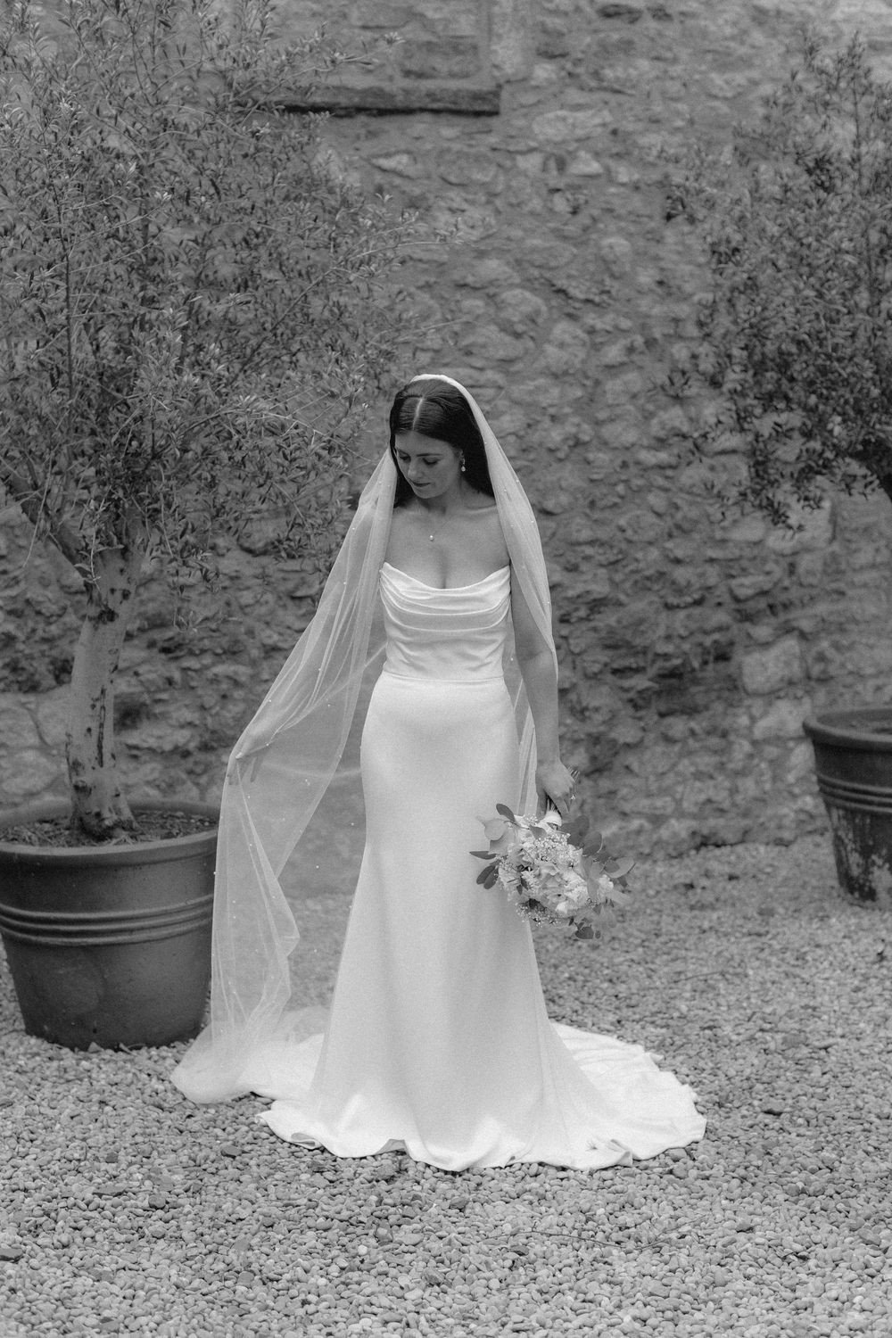 Black and white photo of a bride in a flowing white dress and veil sitting on stone steps by potted plants.