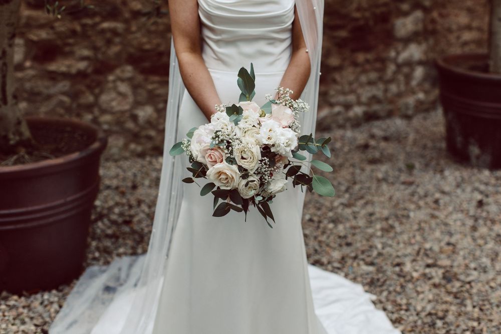 A white wedding bouquet with blush roses and greenery held against a flowing wedding dress.