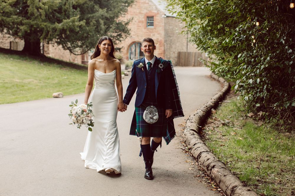 A couple walks together on a stone path surrounded by lush greenery at their outdoor wedding.