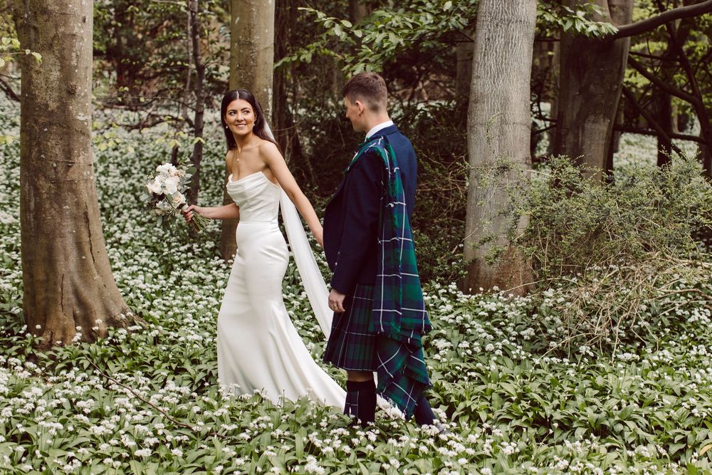 A couple walks together through a woodland setting, with the groom wearing traditional Scottish tartan and bride in white gown.