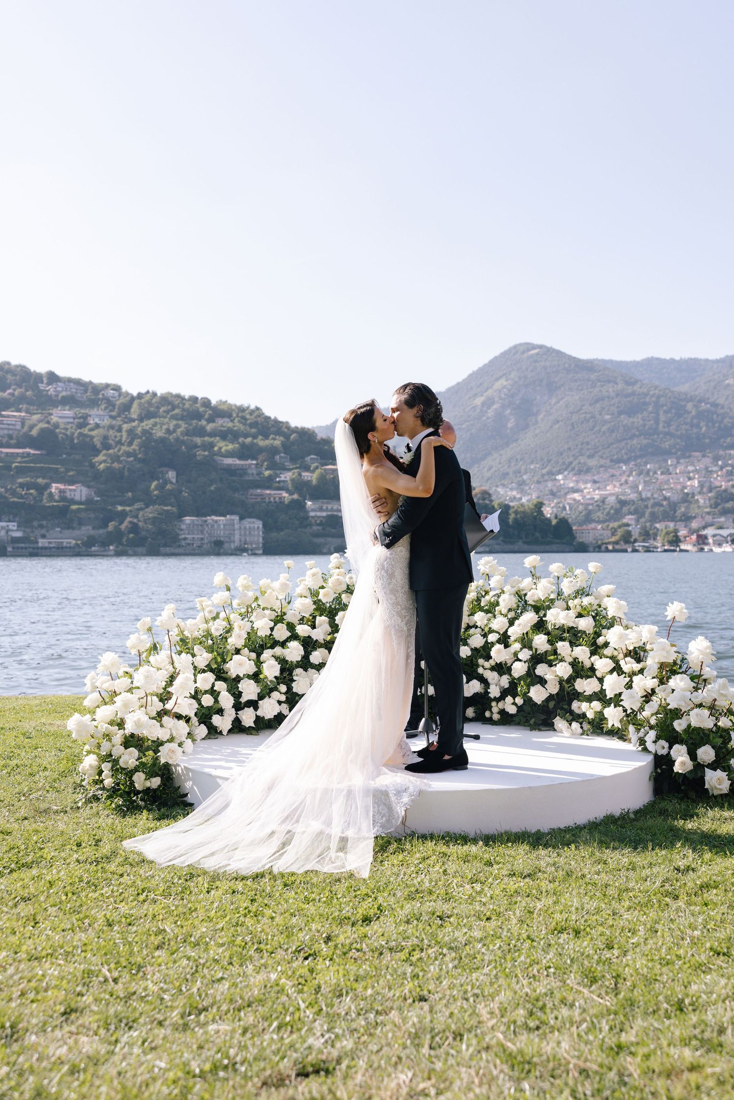 A couple walks down an outdoor aisle lined with flowers and cypress trees leading to an elegant villa entrance.