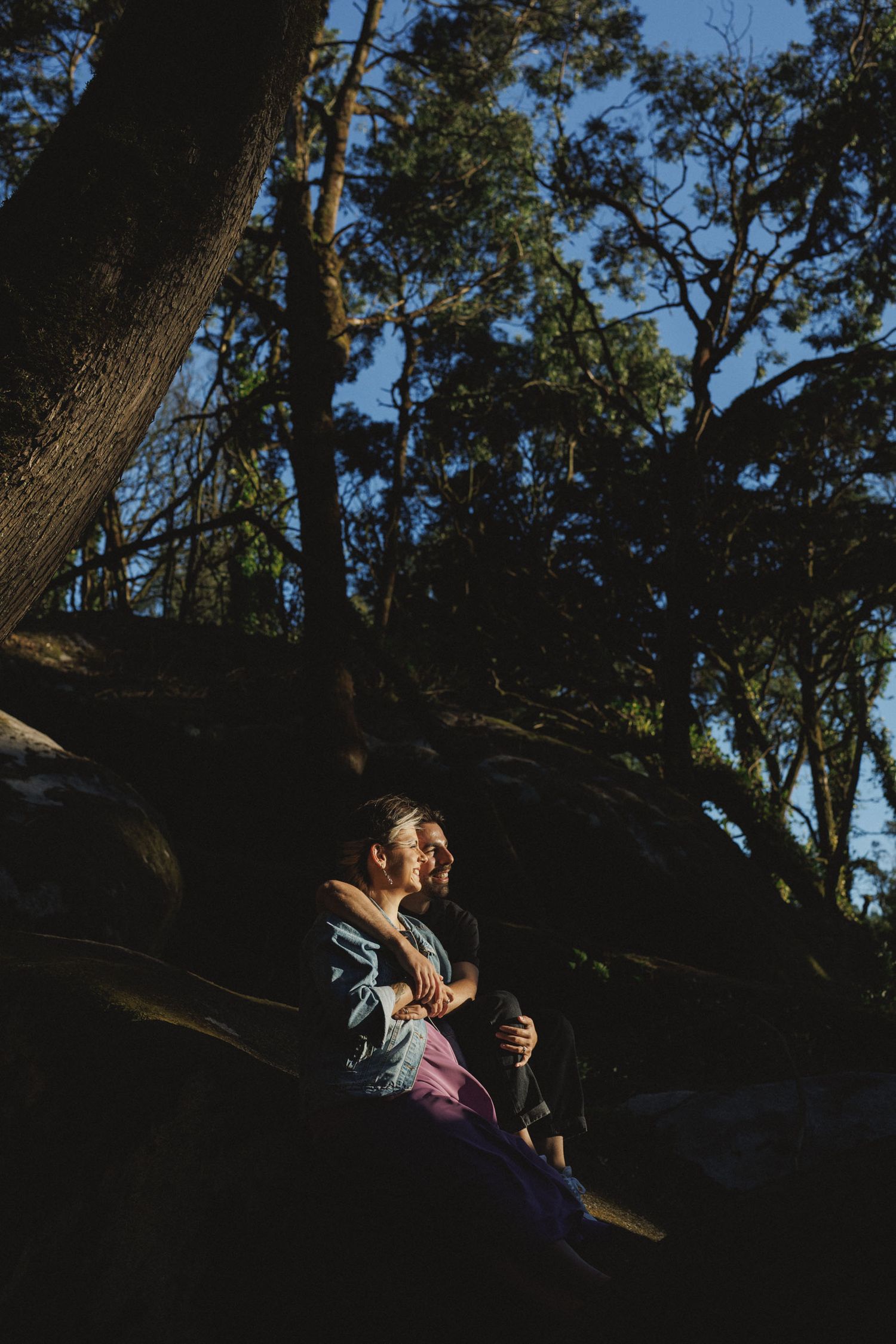 Wide shot of couple sitting on rocks surrounded by trees during sunset engagement session in Sintra, Portugal