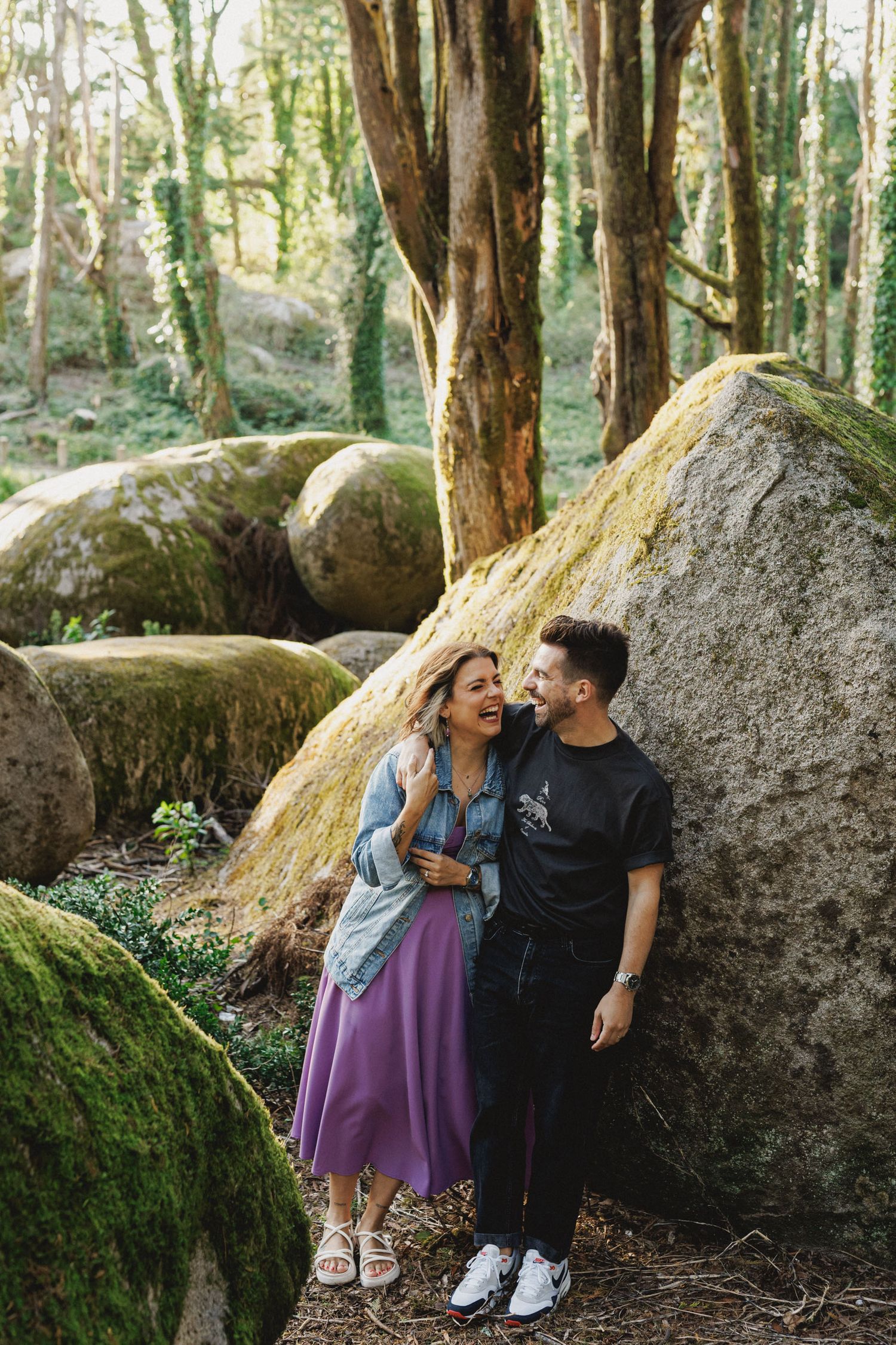 Playful couple standing between large mossy rocks during engagement photoshoot in Sintra forest, Portugal