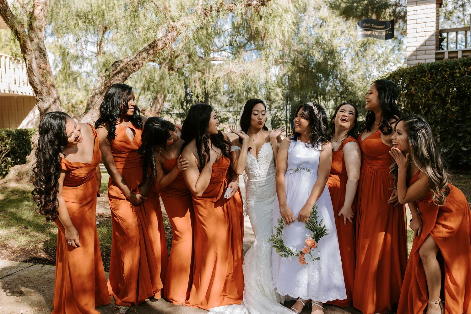 A wedding party poses outdoors wearing burnt orange bridesmaid dresses and a white bridal gown at Rancho Bernardo Inn.