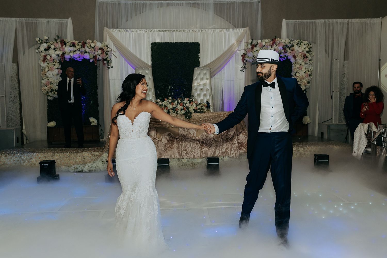 A couple shares their first dance as newlyweds on a white dance floor decorated with elegant floral arrangements at Babylon Palace.