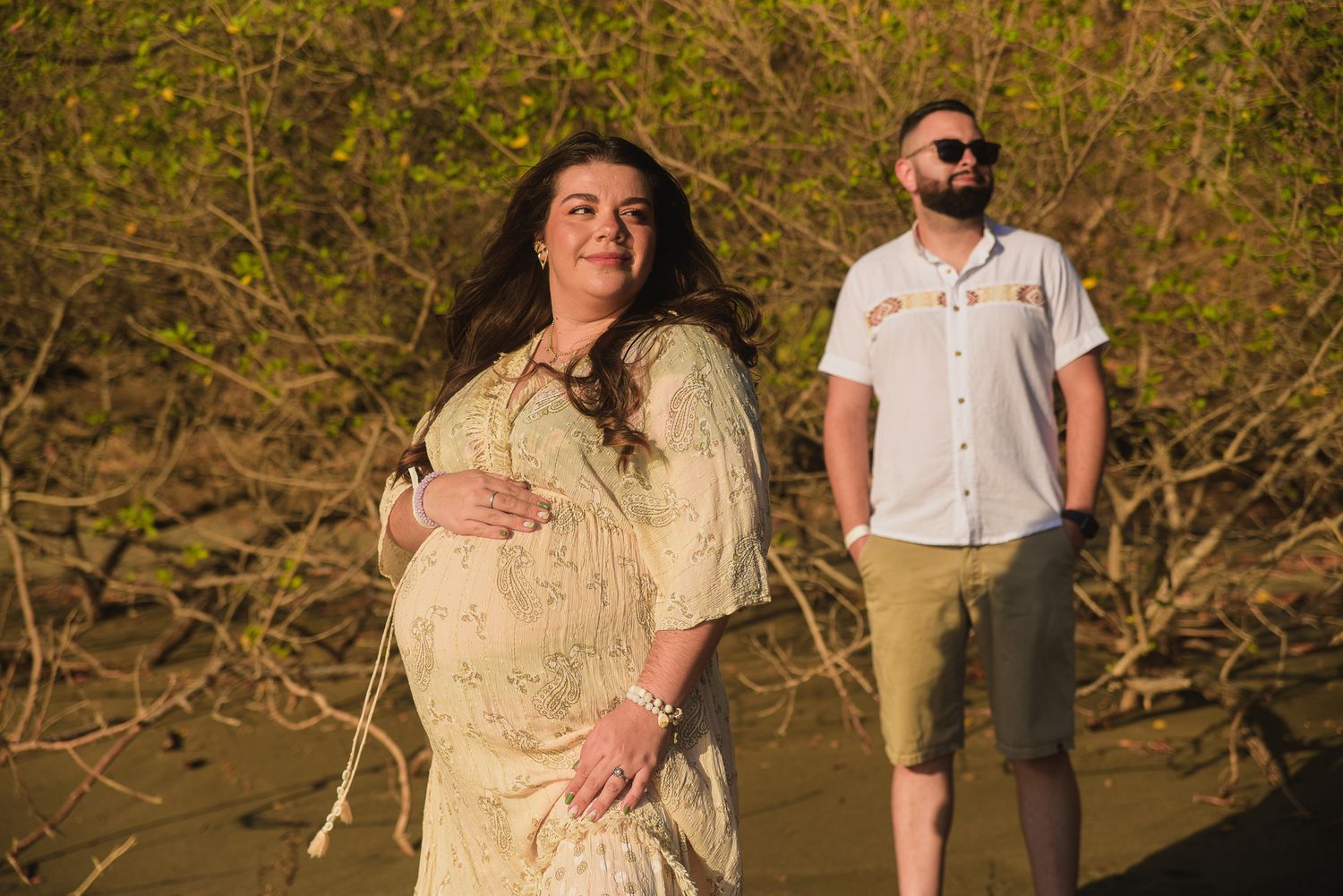 Pregnant woman in flowy dress cradling baby bump outdoors with man standing behind her in golden sunlight at four Season Papagayo