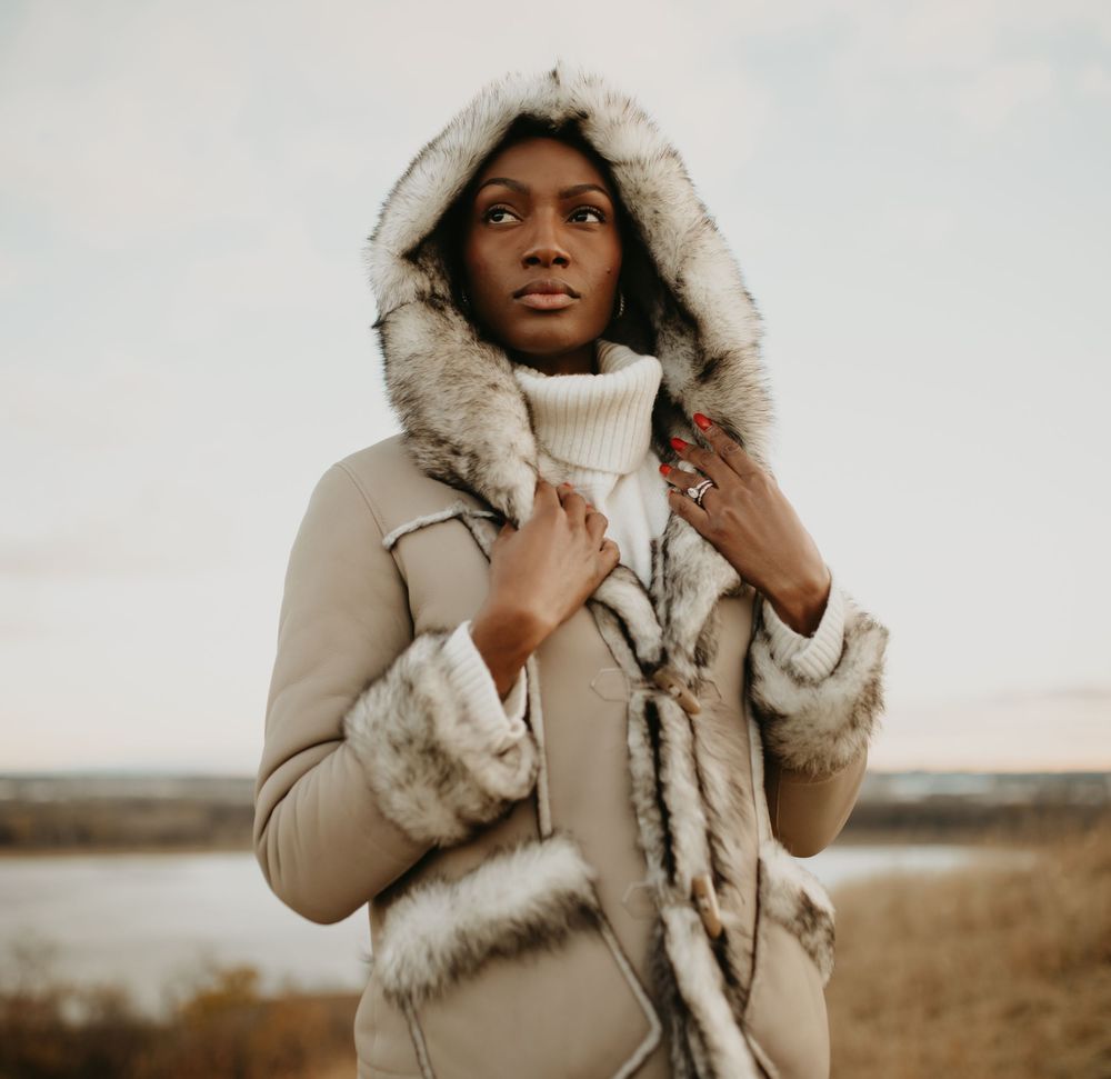 Person wearing a beige winter coat with fur-lined hood stands in golden outdoor field during autumn season.