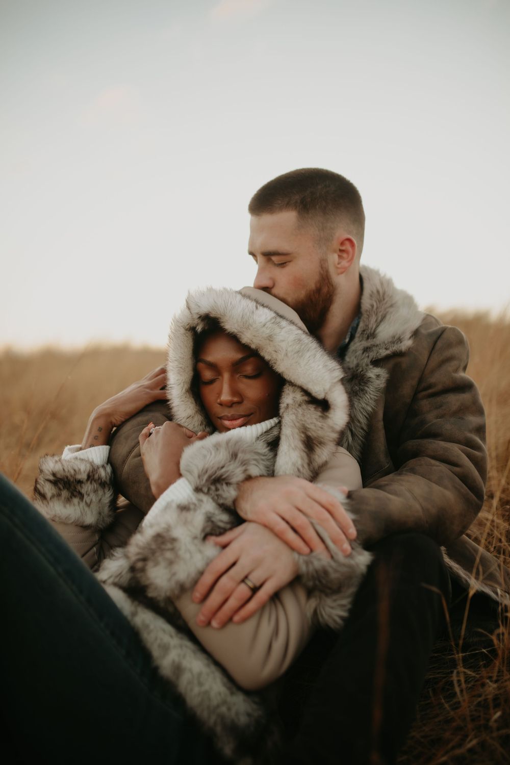 Intimate embrace captured in warm golden light with fur-lined winter coats in a natural outdoor setting.
