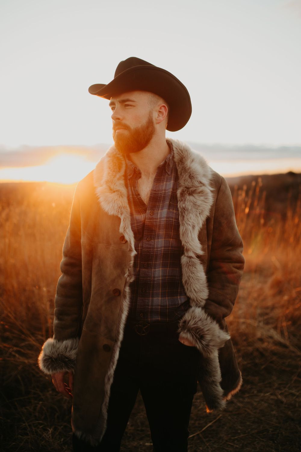 Cowboy in sheepskin jacket stands in golden sunlit field during sunset.