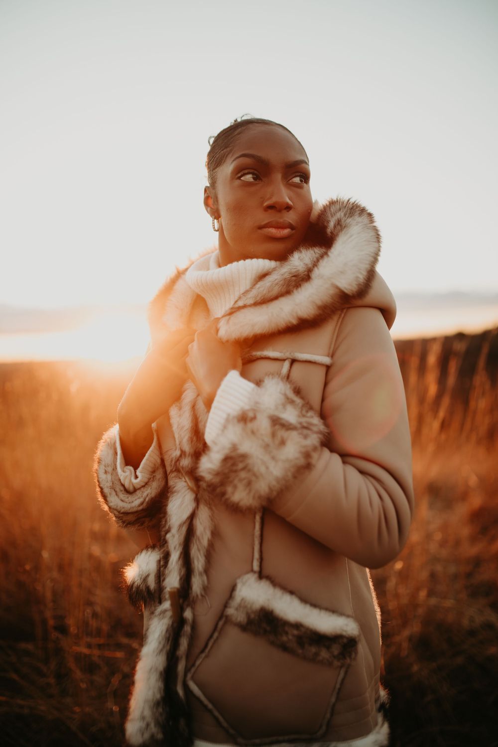 A person wearing a cozy beige winter coat with fur trim stands in warm sunset light against a natural landscape.