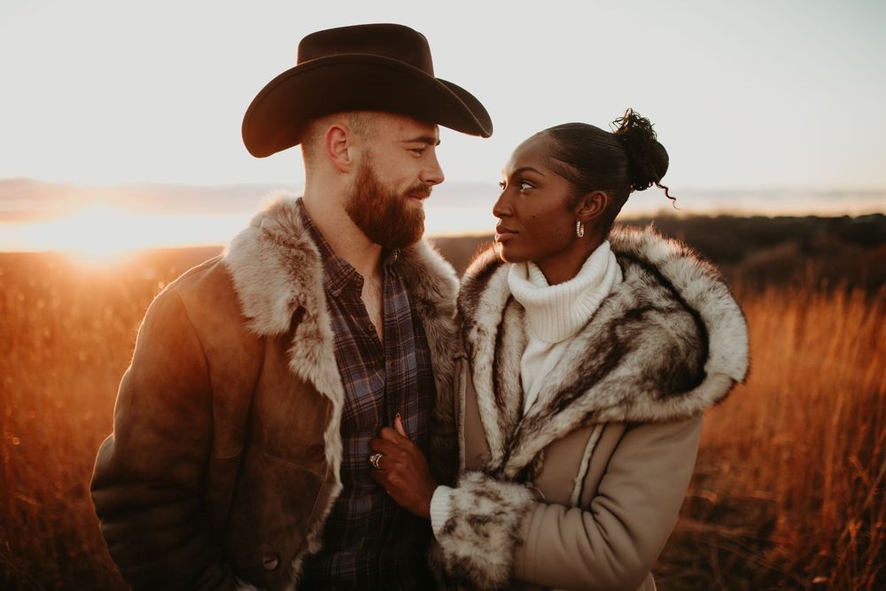 A couple in winter clothing share an intimate conversation in a golden-lit field at sunset.
