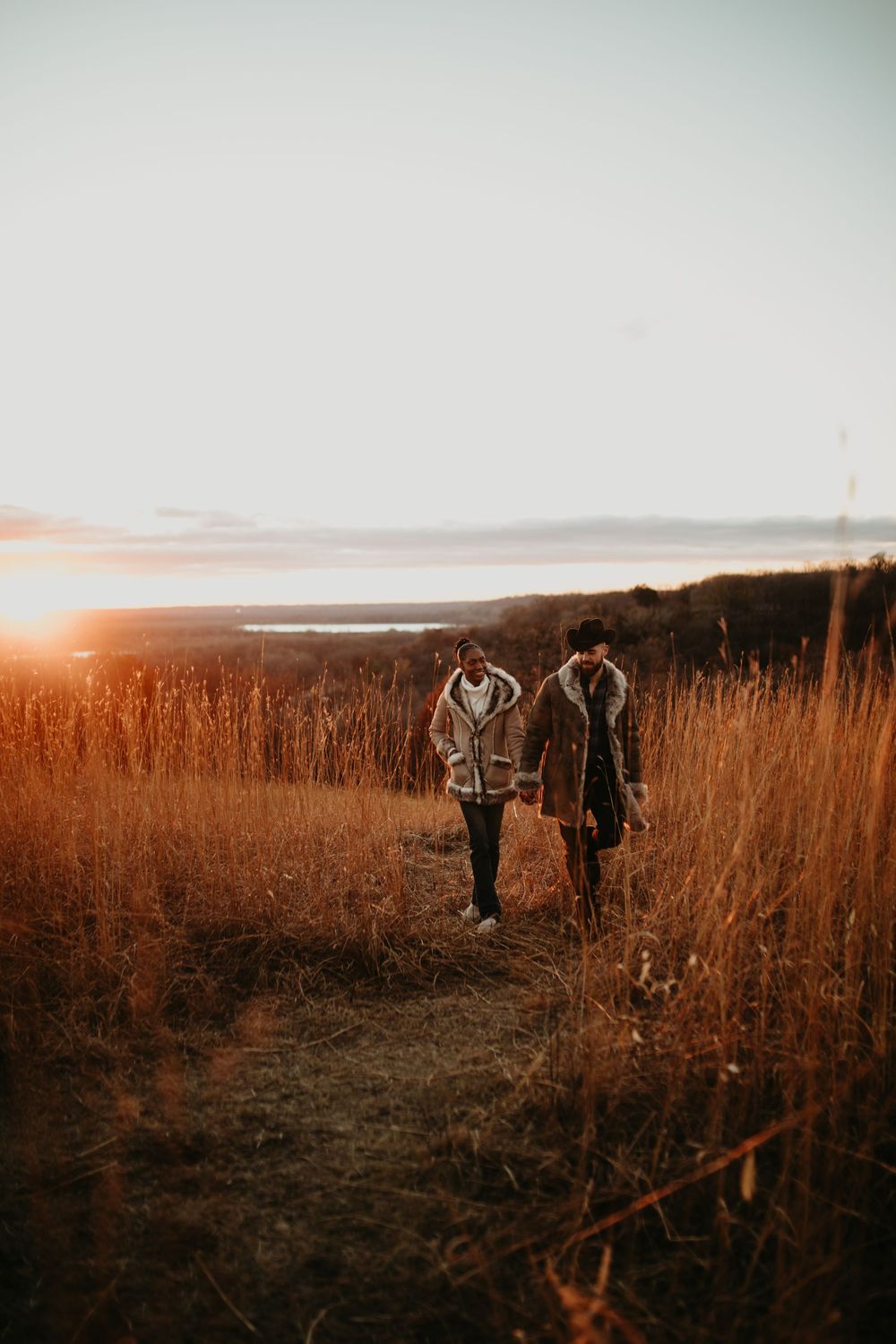 Two figures walking through golden autumn field at sunset with distant water view.