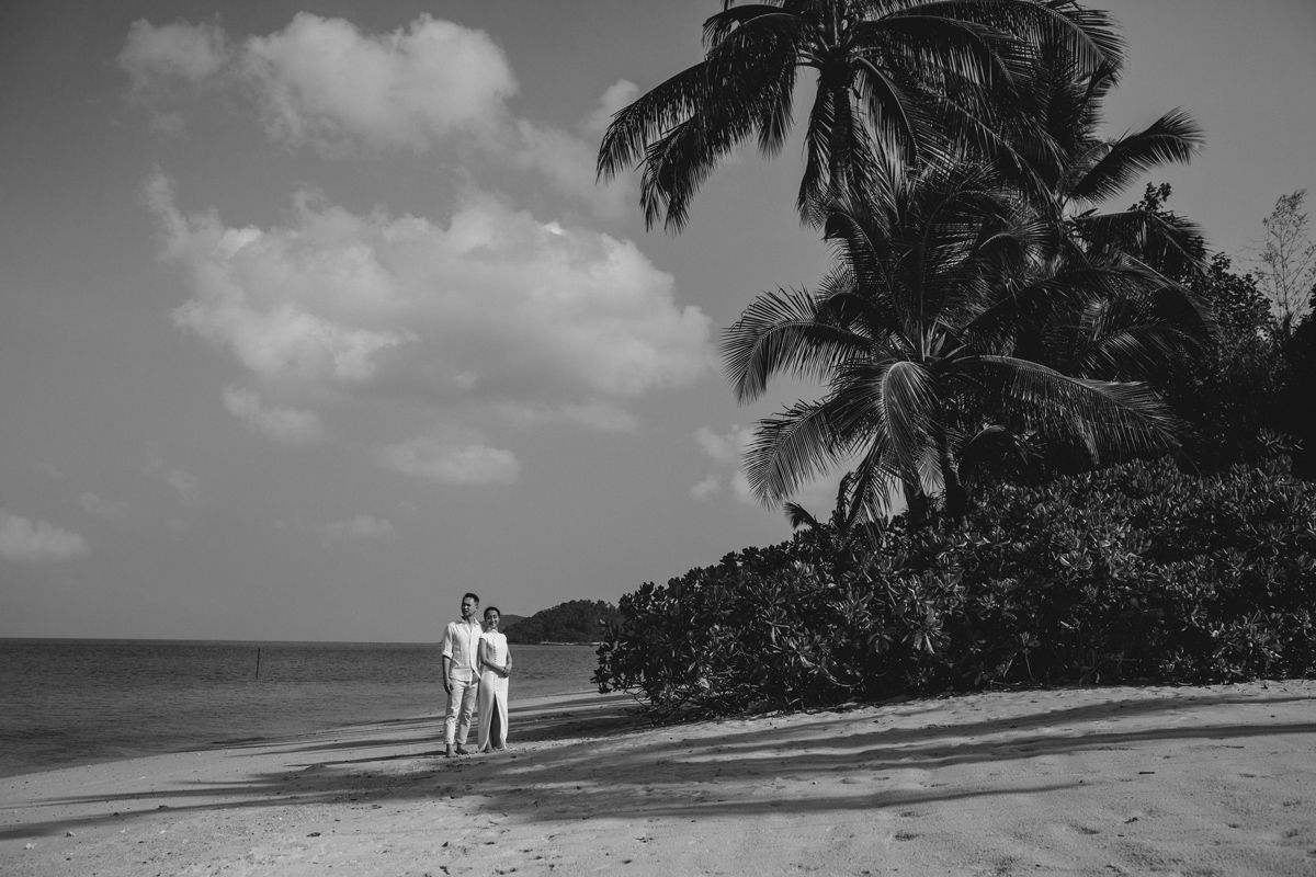 Black and white wide shot of couple walking along private beach in Koh Samui, framed by palm trees and ocean, cinematic destination pre-wed