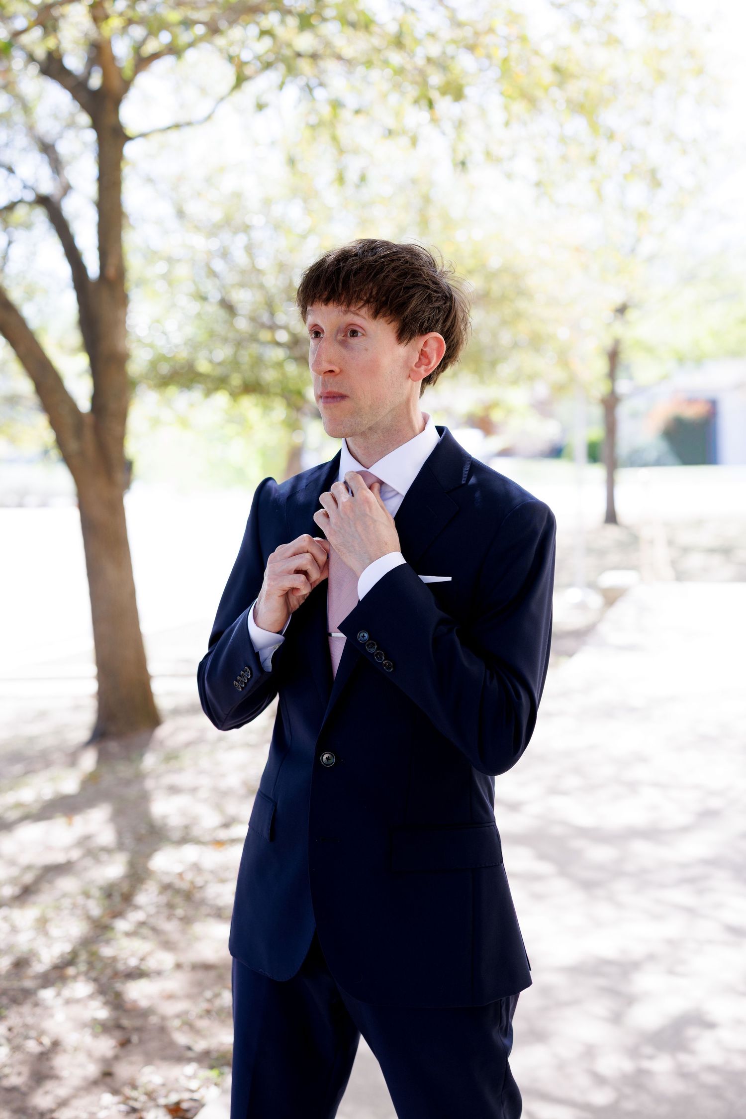 Groom adjusts his tie while standing outdoors in a navy suit.