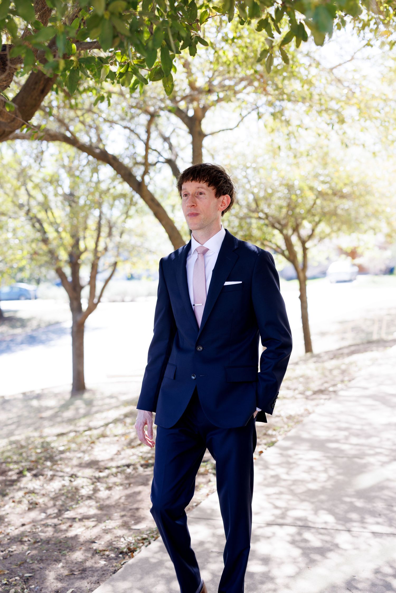 A groom in a navy suit and pink tie poses outdoors among trees on his wedding day.