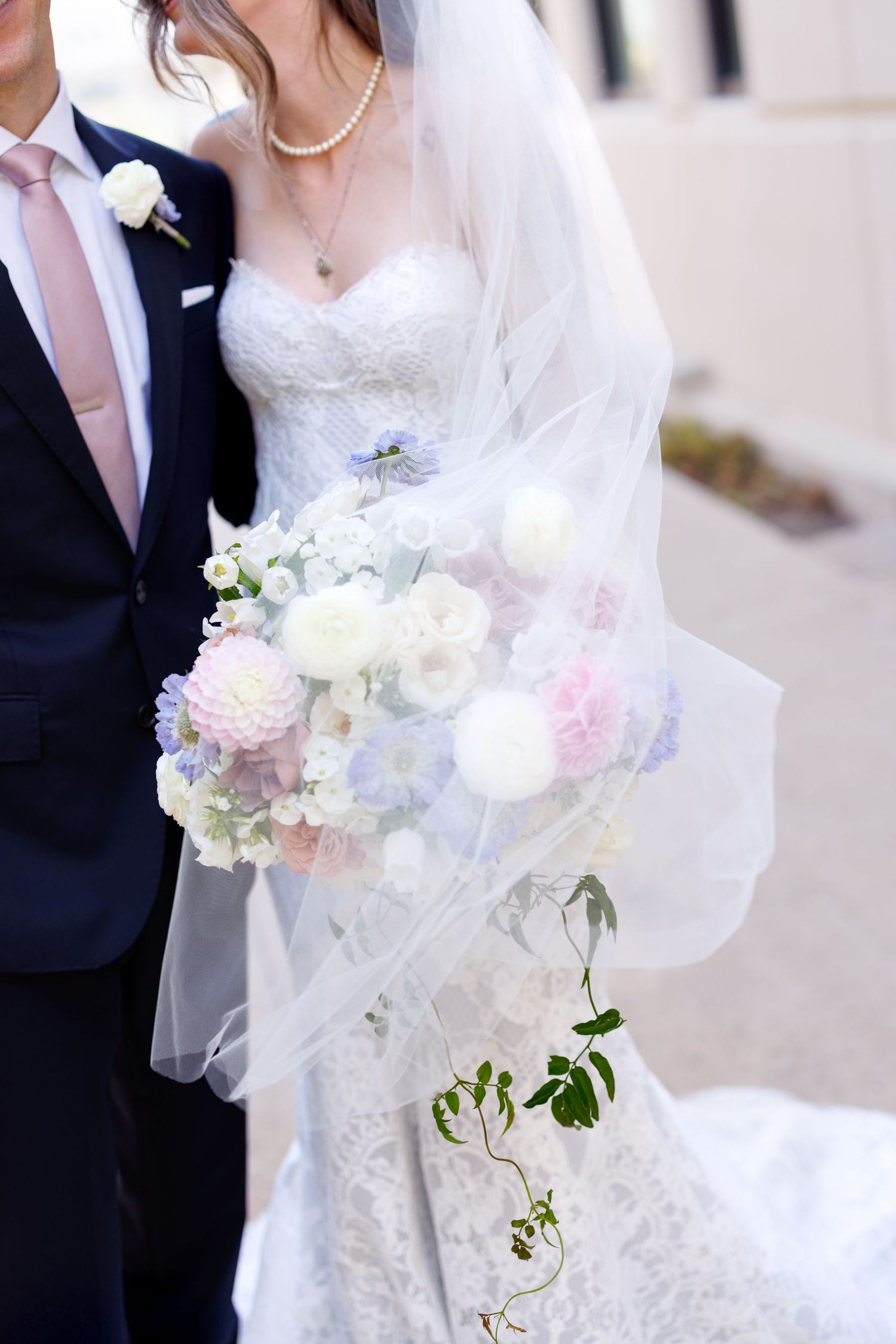 A bride in a white lace gown holds a delicate bouquet of pink and white flowers with pearls.