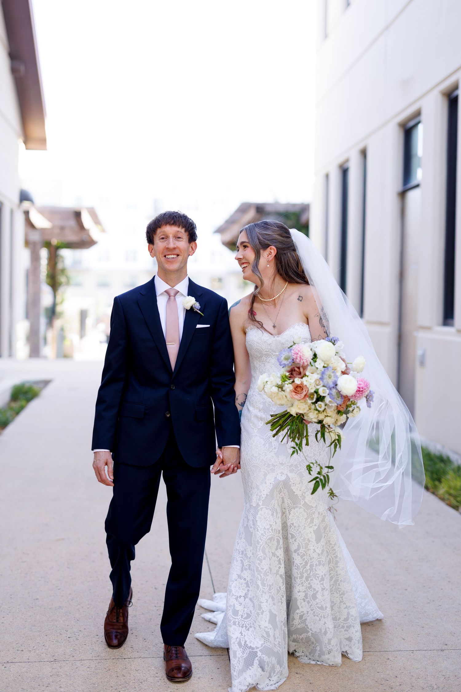 A wedding couple walks together along a white building exterior in formal attire with flowing bridal gown and bouquet.