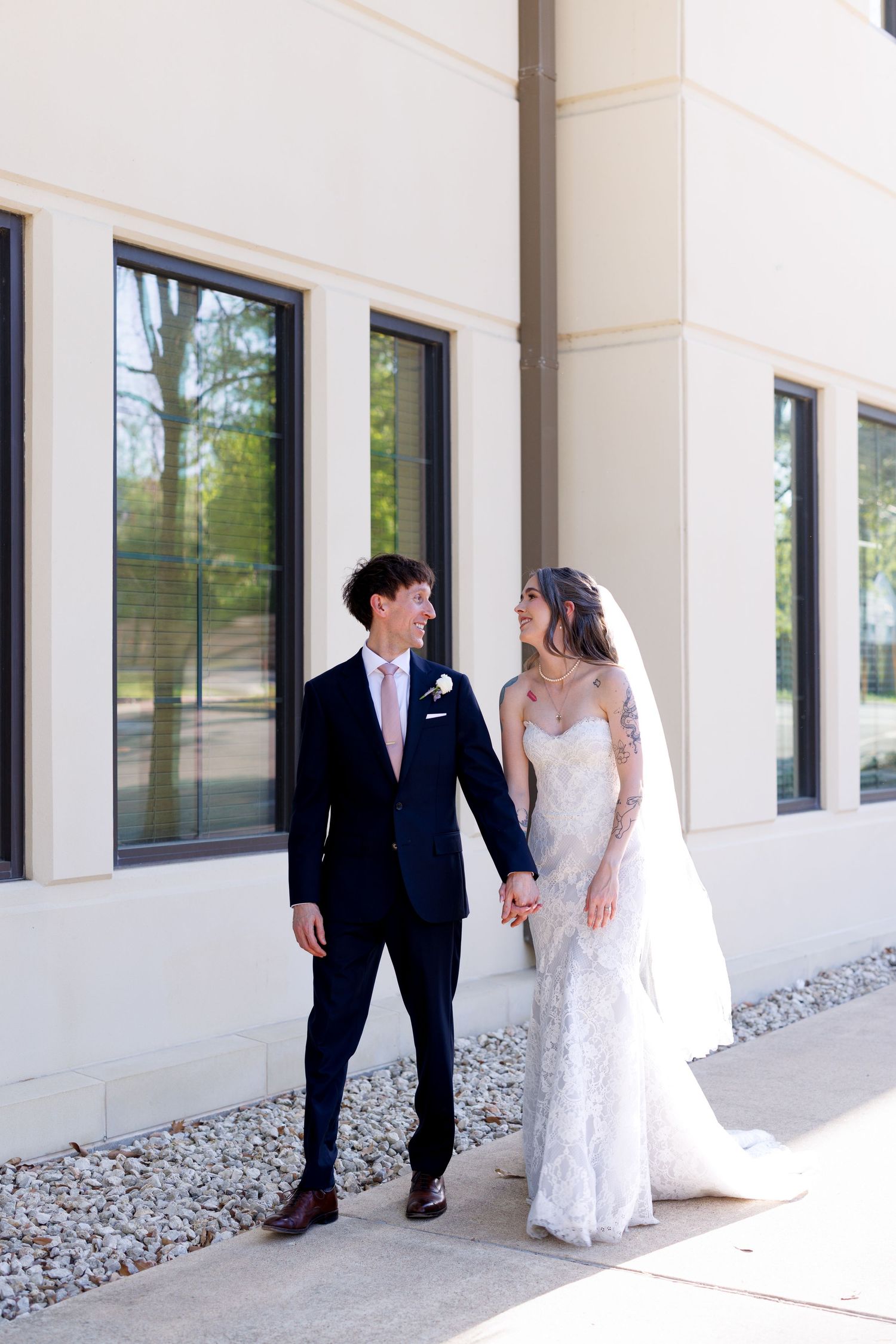 Couple in wedding attire share candid moments along a modern building exterior.