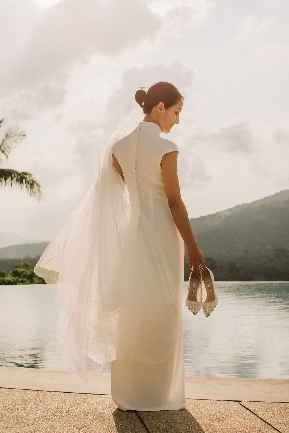 Bride in elegant dress walking barefoot by infinity pool at InterContinental Koh Samui, holding shoes, soft sunrise light over tropical hill