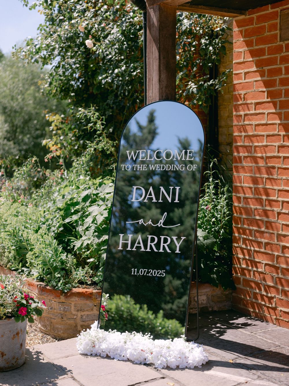 A series of elegant glass welcome signs with white text displayed against brick walls in an outdoor garden setting.