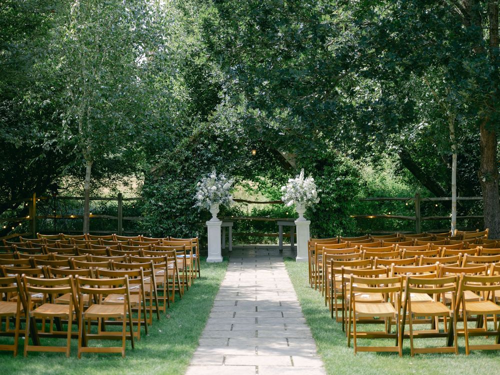 Rows of wooden folding chairs arranged on a green lawn for an outdoor wedding ceremony beneath towering trees.