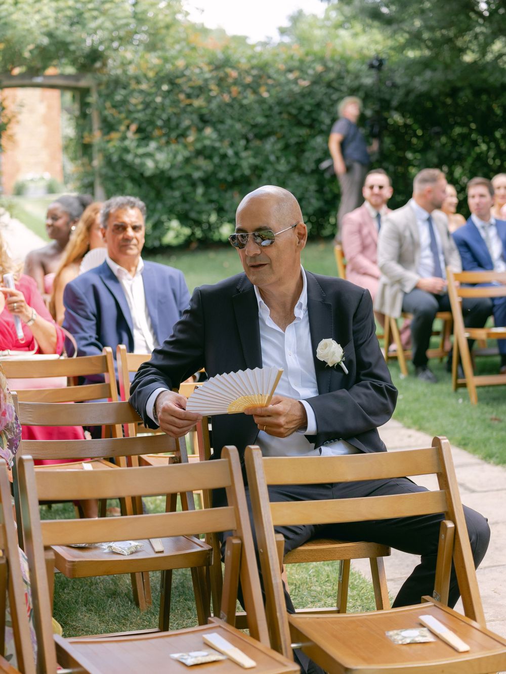 Wedding guests seated on wooden folding chairs at an outdoor garden ceremony with one attendee holding a paper fan.