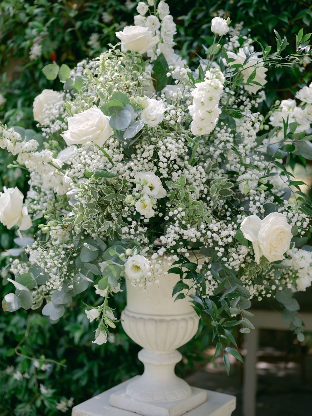 Elegant white floral arrangement with roses and baby's breath in a stone urn against lush greenery.