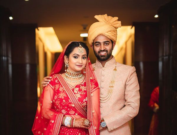 A newlywed couple in traditional Indian wedding attire poses in an elegant indoor setting.