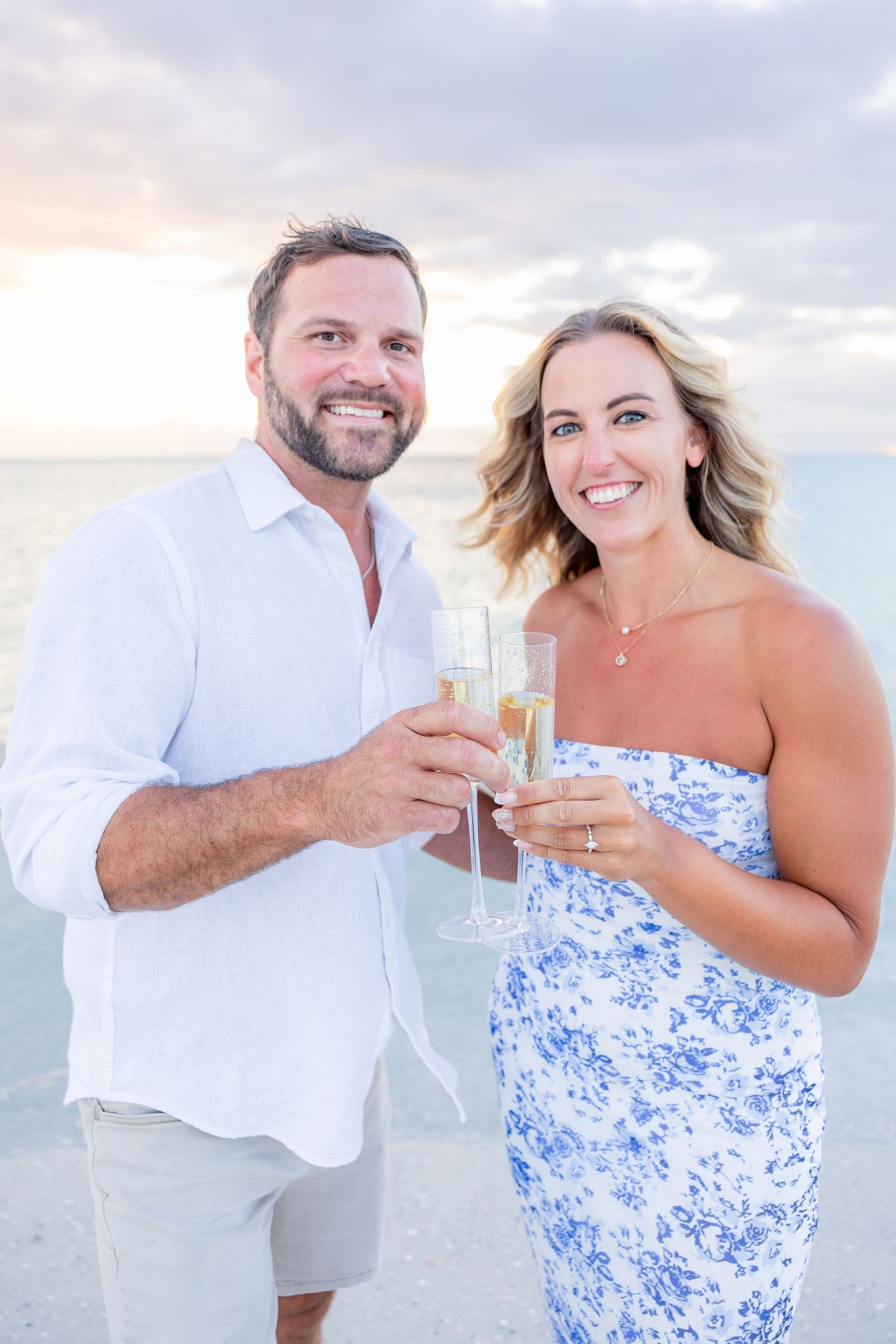 Smiling couple toasting with champagne glasses on beach at sunset in coordinating outfits.