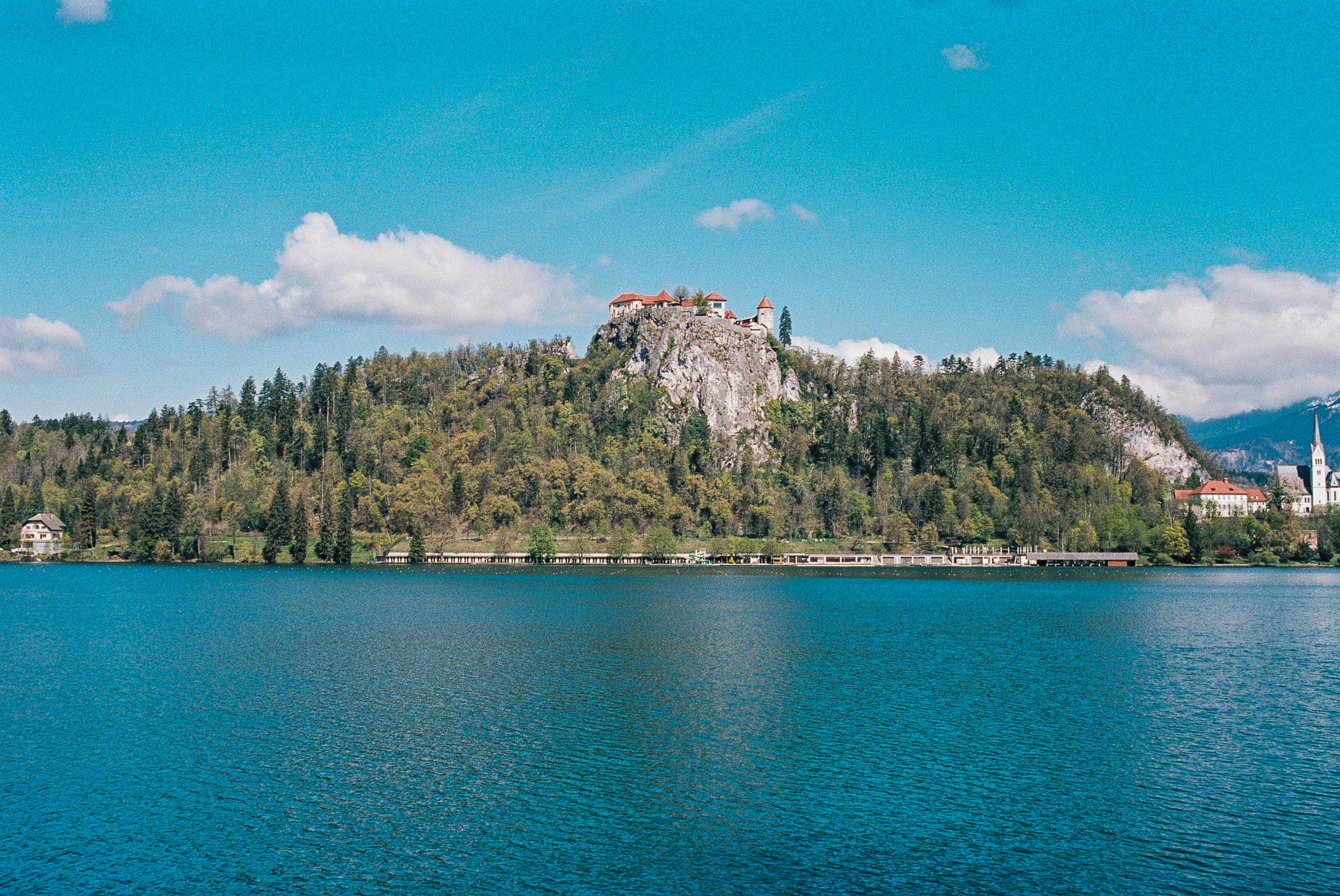 Rocky cliff rising above Lake Bled with dense forest and mountains in background.