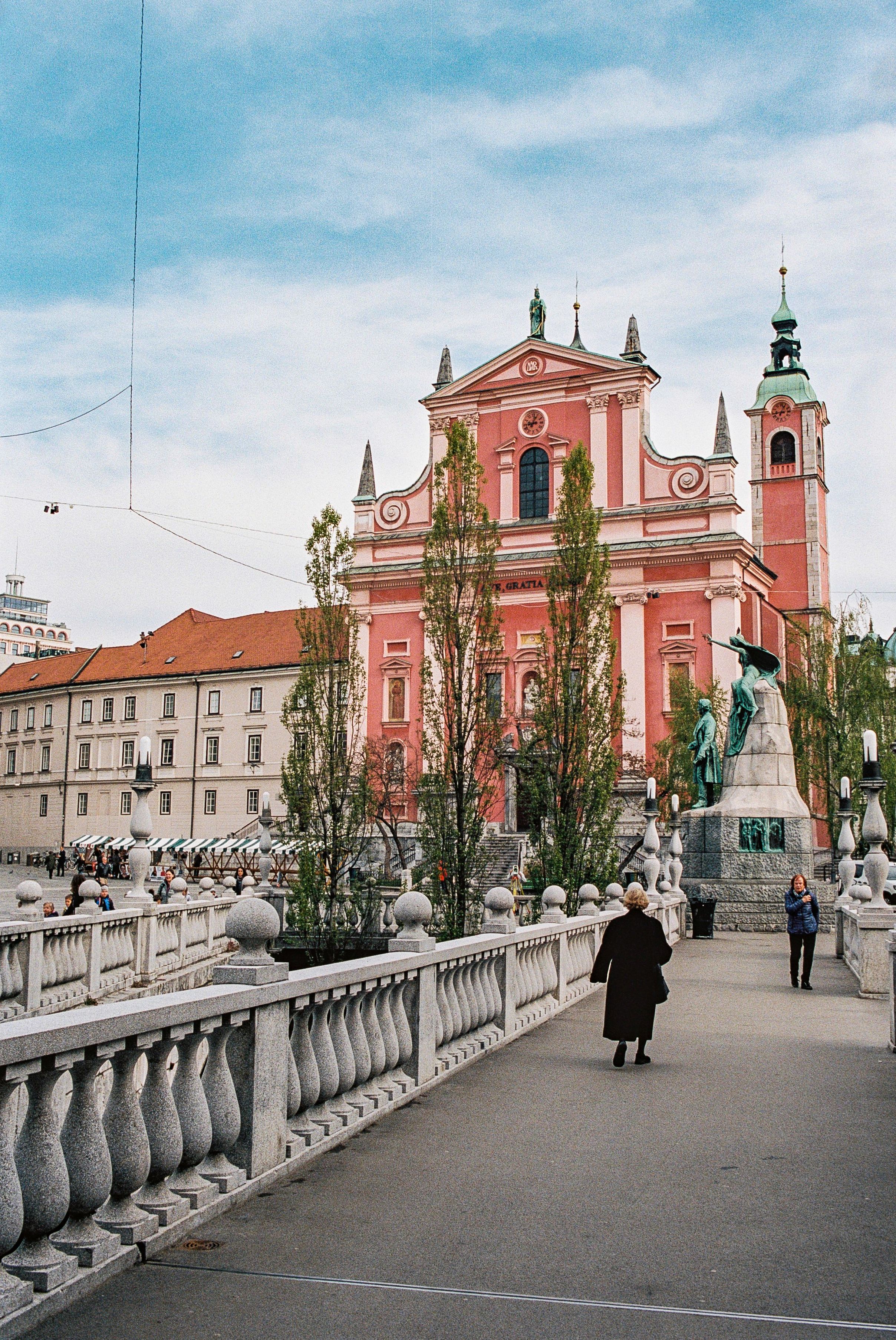 Pink Baroque church with twin towers and white balustrade bridge in Ljubljana's city center.