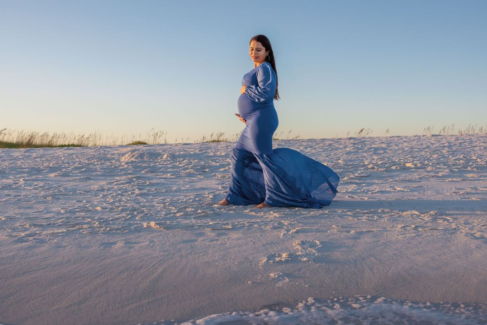 A pregnant woman in a flowing blue dress poses on a white sandy beach during golden hour.