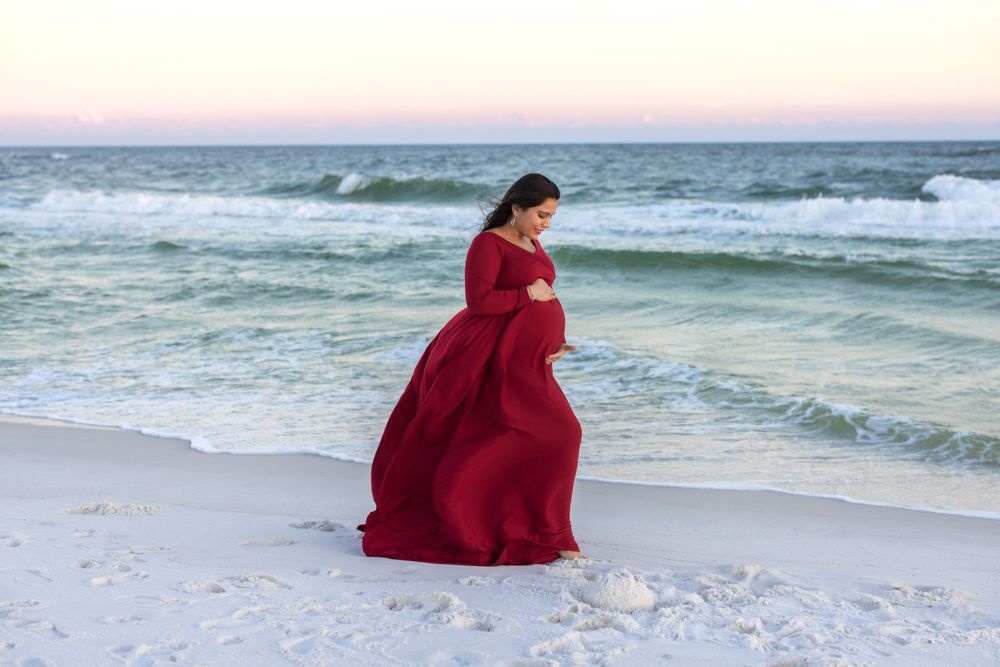 A pregnant woman in a red flowing dress walks along the shoreline at sunset.