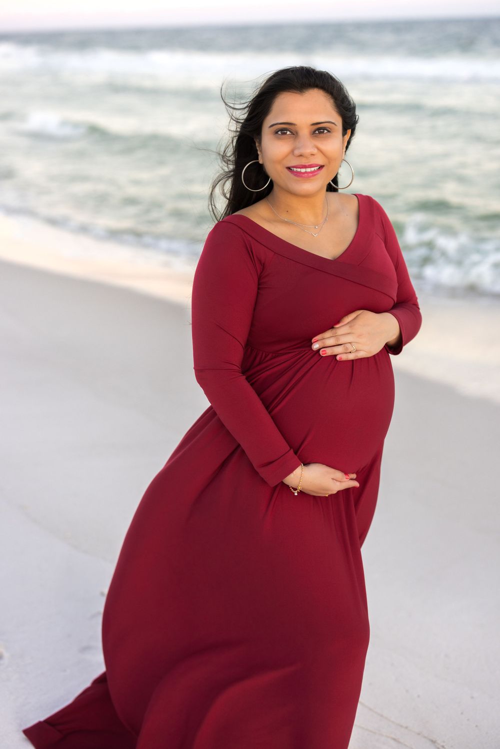A pregnant woman in a flowy burgundy maxi dress poses on a beach with waves in the background.