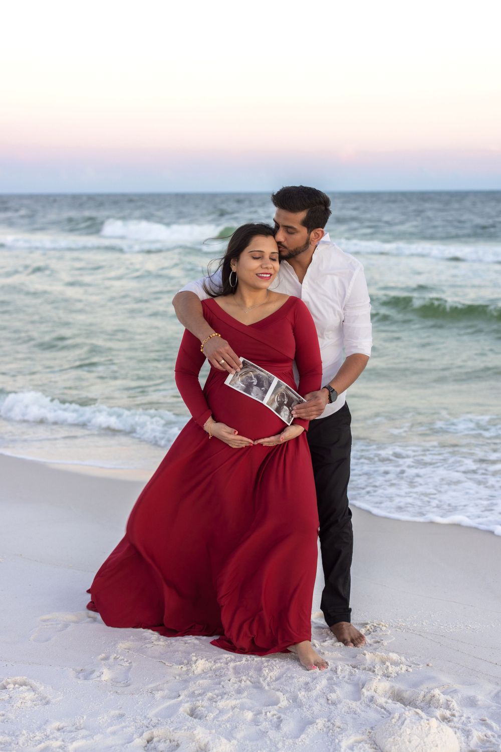 A couple embraces on a beach at sunset during a maternity shoot featuring a flowing red dress.