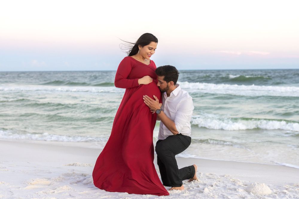 A romantic maternity moment captured on a white sandy beach with waves in the background.