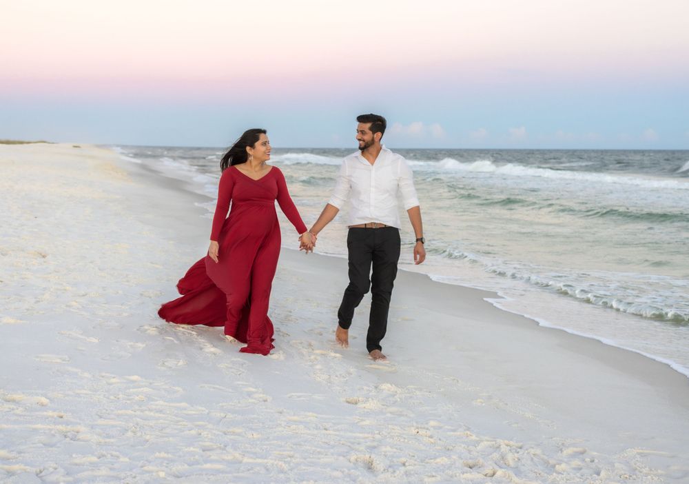 A couple walks hand in hand on a white sandy beach at sunset, with the woman wearing a flowing red dress.
