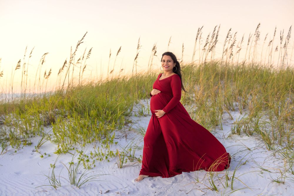 A pregnant woman in a flowing red dress poses among beach grass during golden hour.